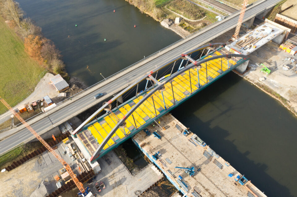 Luftaufnahme einer Brücke im Bau mit gelbem Deck, die von zwei großen Bögen getragen wird und über einen Fluss führt. Auf beiden Seiten der Brücke befinden sich Baustellen mit Kränen und Baugeräten, ein Auto fährt über die Brücke.