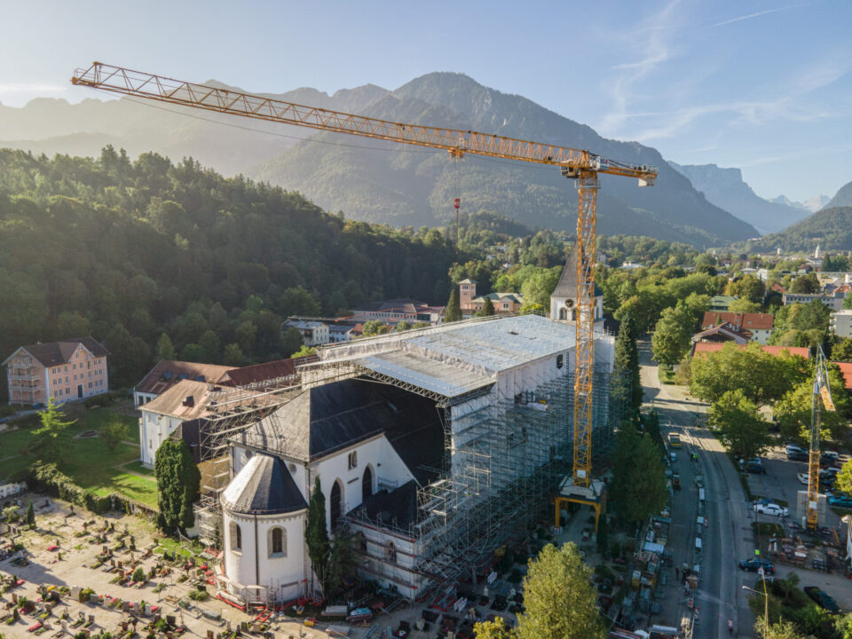 Das Bild zeigt eine große Baustelle, auf der eine Kirche renoviert wird. Die Kirche ist vollständig von Gerüsten umgeben, und zwei hohe Baukräne stehen links und rechts daneben. Im Hintergrund sind bewaldete Berge und verstreute Gebäude zu sehen, bei klarem Himmel mit wenigen Wolken.