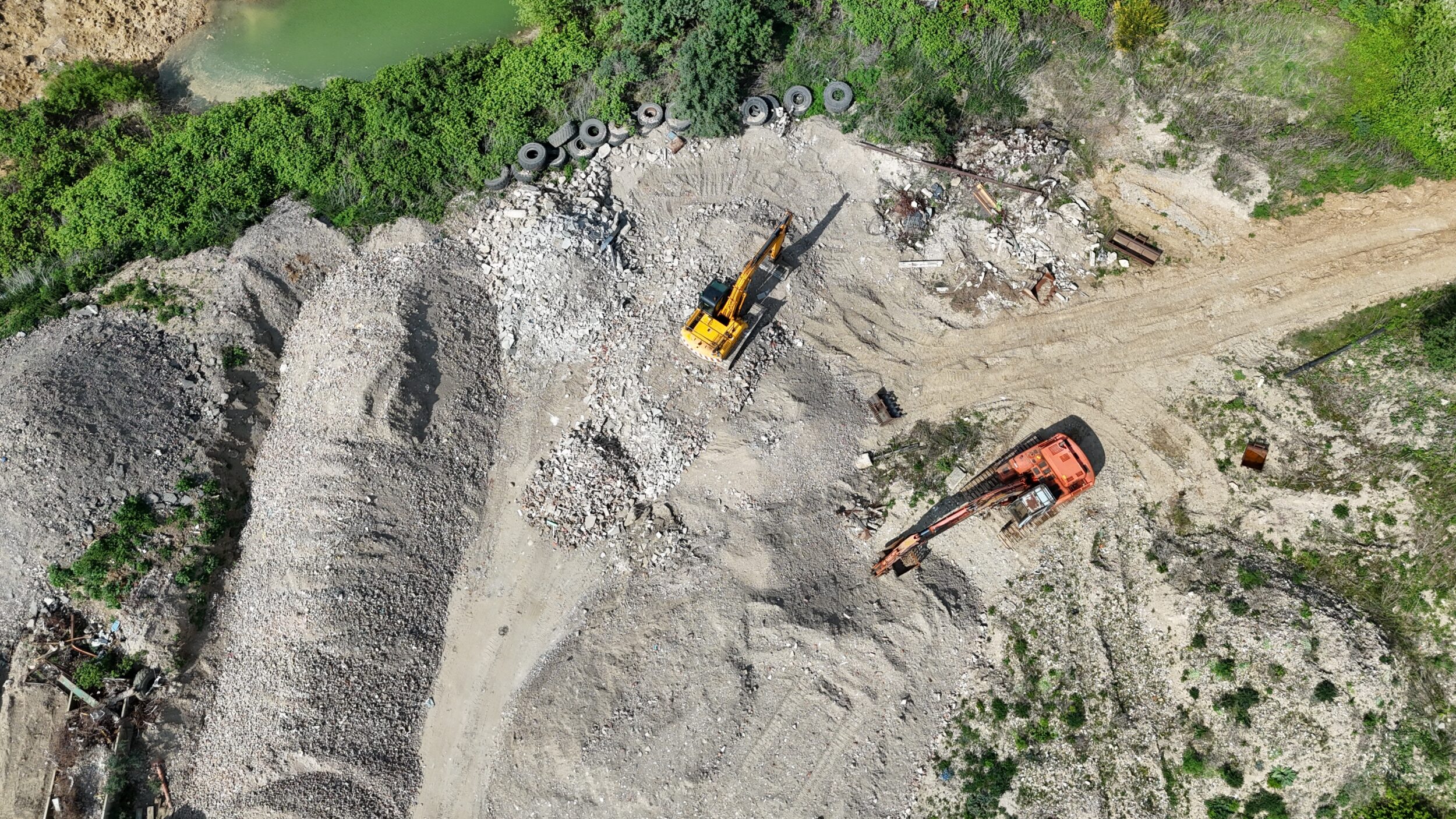 Das Bild zeigt eine Baustelle aus der Vogelperspektive mit zwei großen Baggern, die auf einer Fläche aus Schutt und Erde arbeiten. Neben den Maschinen liegen große Haufen aus Steinen und Baumaterial, während im oberen Bereich grüne Vegetation und ein kleiner Wasserbereich zu sehen sind. Mehrere alte Reifen sind am Rand der Baustelle abgelegt.