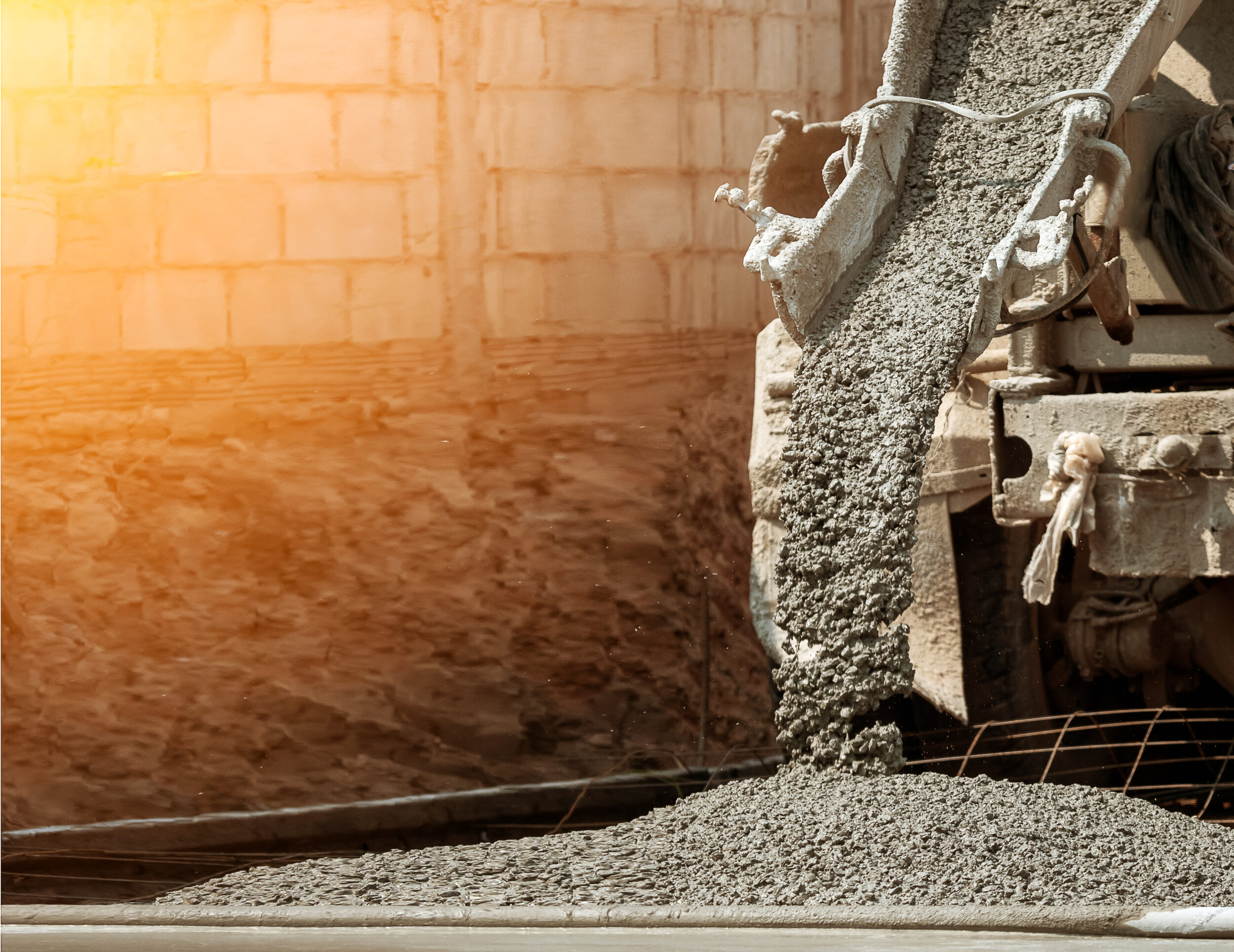 Das Bild zeigt einen Betonmischer, aus dessen Rutsche frischer Beton auf eine vorbereitete Fläche gegossen wird. Der Beton ist grobkörnig und verteilt sich auf einem Untergrund, der mit einer Metallarmierung versehen ist. Im Hintergrund sind eine Mauer aus hellen Betonblöcken und warmes Sonnenlicht zu sehen, das die Szene beleuchtet.