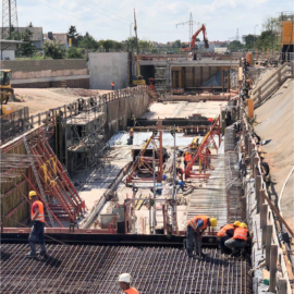 Das Bild zeigt eine große Baustelle für den U-Bahn-Bau, aufgenommen bei Tageslicht. Im Vordergrund arbeiten mehrere Personen in orangefarbenen Sicherheitswesten auf einer Fläche mit dicht verlegtem Bewehrungsstahl. Dahinter sind rote Stahlträger und Schalungselemente zu sehen, die den Tunnelbereich stützen. Im Hintergrund befinden sich weitere Baugeräte, darunter ein Bagger und ein Kran, sowie Betonwände, die den Bauabschnitt begrenzen. Strommasten und ein blauer Himmel mit vereinzelten Wolken runden die Szene ab.