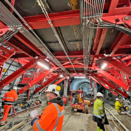 Das Bild zeigt den Innenbereich eines U-Bahn-Tunnels im Bau, der von einer massiven roten Stahlkonstruktion gestützt wird. Die Konstruktion besteht aus zahlreichen Verstrebungen und Trägern, die den Tunnel stabilisieren, während Kabel und Leitungen ordentlich entlang der Decke verlaufen. Mehrere Personen in Sicherheitskleidung und Helmen arbeiten im Tunnel, und im Hintergrund ist ein kleiner Baustellenfahrzeug zu sehen. Die Szene ist gut ausgeleuchtet durch mehrere helle Lampen, die an der Konstruktion befestigt sind.