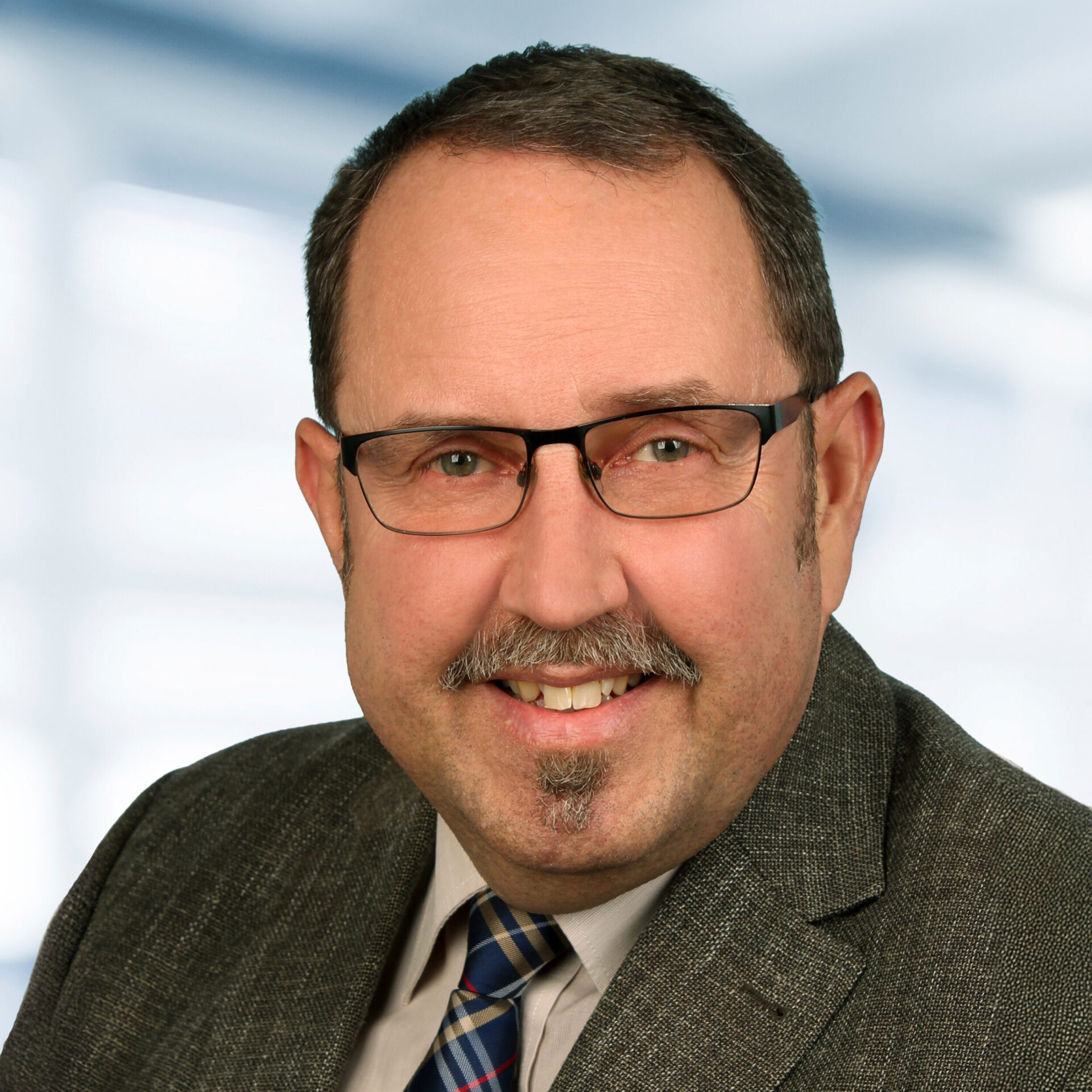 Auf dem Bild ist ein Mann in einem professionellen Porträt zu sehen, der freundlich in die Kamera lächelt. Er trägt eine Brille, ein Jackett mit Hemd und Krawatte, der Hintergrund ist hell und unscharf, sodass die Person klar im Vordergrund steht.