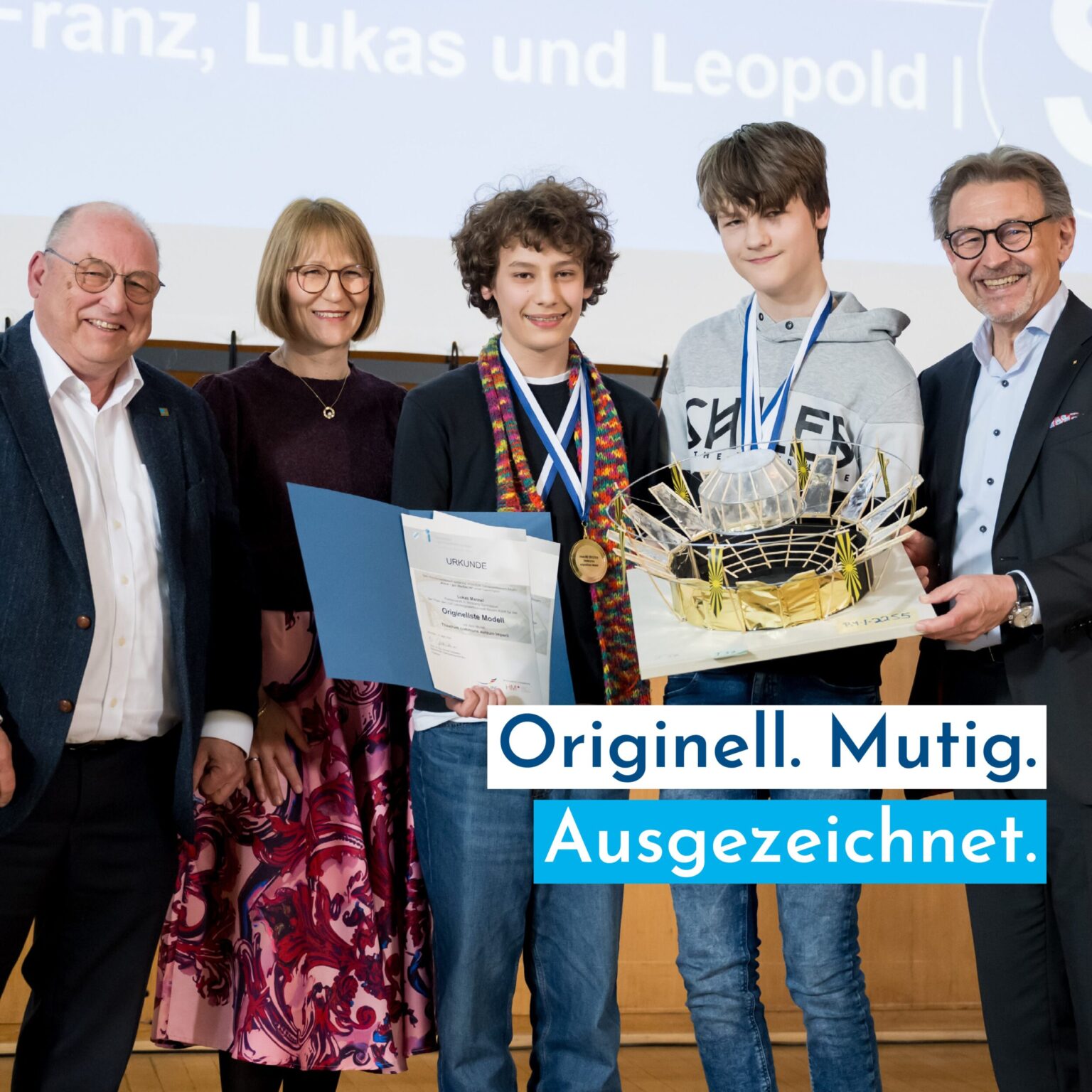 Auf dem Bild steht eine Gruppe von fünf Personen auf einer Bühne und blickt lächelnd in die Kamera. Zwei Jugendliche in der Mitte tragen Medaillen, einer hält eine Urkunde und der andere ein Modell, während die Erwachsenen danebenstehen. Im Vordergrund befindet sich ein großer Schriftzug, der auf eine Auszeichnung hinweist.