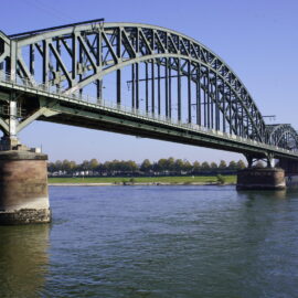 Auf dem Bild ist eine große Stahlbrücke mit mehreren Bögen zu sehen, die über einen breiten Fluss führt. Die Brücke ruht auf massiven Pfeilern und verbindet zwei Ufer mit einer flachen Landschaft im Hintergrund. Das Wasser fließt ruhig unter der Konstruktion hindurch und spiegelt Teile der Brücke wider.