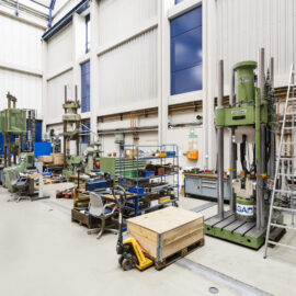 Auf dem Bild ist eine große Industriehalle mit verschiedenen Maschinen, Werkbänken und technischen Anlagen zu sehen. Mehrere Arbeitsplätze mit Werkzeugen, Materialwagen und Lagerflächen sind über die Halle verteilt. Die hohe Decke und die großzügige Raumaufteilung vermitteln den Eindruck einer Werkstatt oder eines Prüflabors für technische Arbeiten.