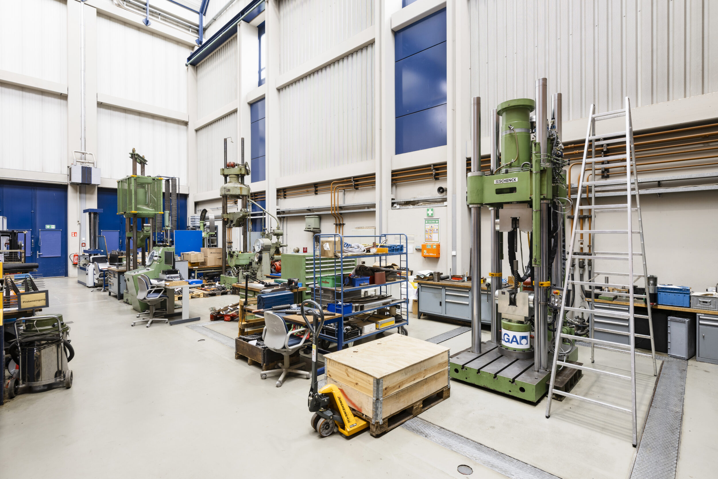 Auf dem Bild ist eine große Industriehalle mit verschiedenen Maschinen, Werkbänken und technischen Anlagen zu sehen. Mehrere Arbeitsplätze mit Werkzeugen, Materialwagen und Lagerflächen sind über die Halle verteilt. Die hohe Decke und die großzügige Raumaufteilung vermitteln den Eindruck einer Werkstatt oder eines Prüflabors für technische Arbeiten.