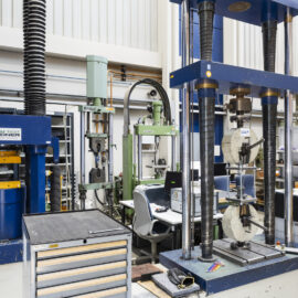 Auf dem Bild sind mehrere große Materialprüfmaschinen in einer Labor- oder Werkstatthalle zu sehen. Die unterschiedlichen Anlagen sind mit Mess- und Prüfgeräten ausgestattet und dienen der Untersuchung mechanischer Eigenschaften von Werkstoffen. Arbeitsplätze, Computer und technische Einrichtungen ergänzen die professionelle Prüf- und Versuchsumgebung.