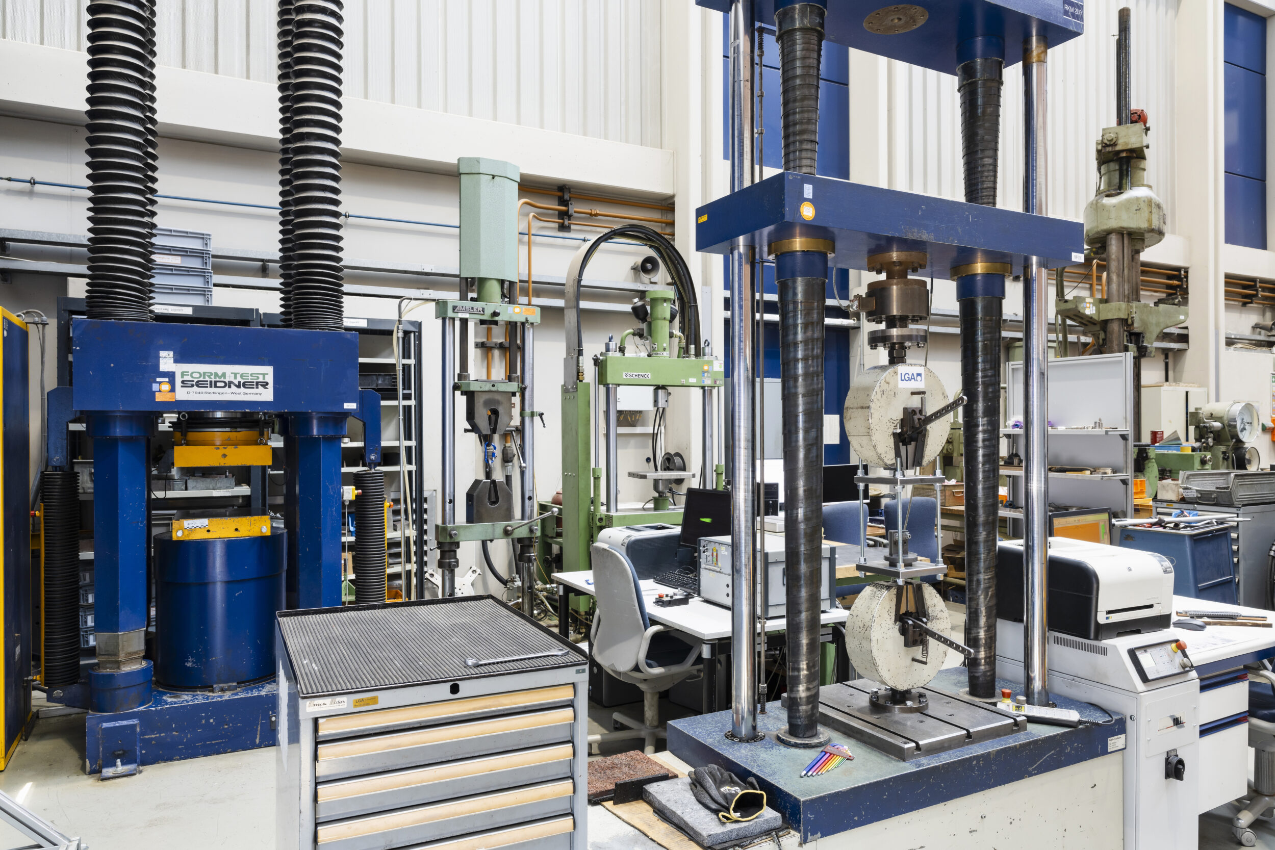 Auf dem Bild sind mehrere große Materialprüfmaschinen in einer Labor- oder Werkstatthalle zu sehen. Die unterschiedlichen Anlagen sind mit Mess- und Prüfgeräten ausgestattet und dienen der Untersuchung mechanischer Eigenschaften von Werkstoffen. Arbeitsplätze, Computer und technische Einrichtungen ergänzen die professionelle Prüf- und Versuchsumgebung.