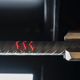 Auf dem Bild ist ein Betonstahlstab in einer Prüf- oder Bearbeitungsvorrichtung eingespannt. Rote Lichtmarkierungen auf der Oberfläche deuten auf eine technische Messung oder Materialuntersuchung hin. Die Aufnahme konzentriert sich auf den Prüfbereich und hebt die Struktur des gerippten Betonstahls deutlich hervor.