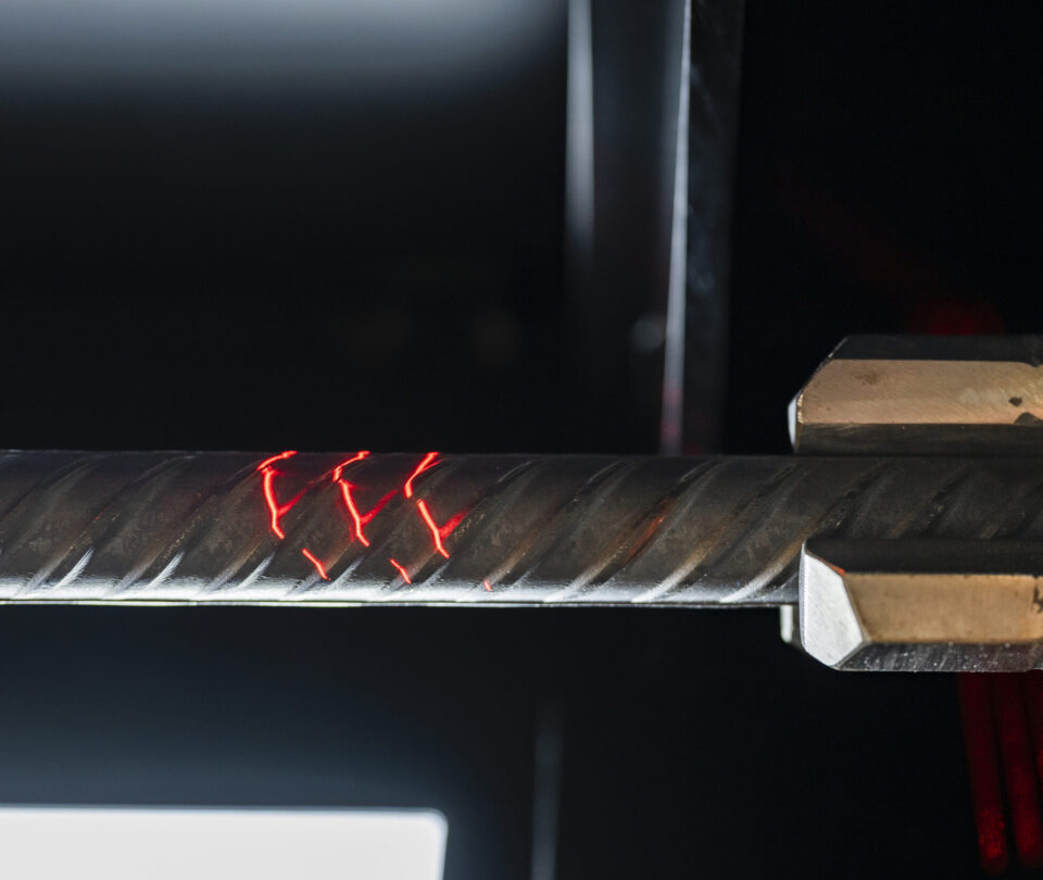 Auf dem Bild ist ein Betonstahlstab in einer Prüf- oder Bearbeitungsvorrichtung eingespannt. Rote Lichtmarkierungen auf der Oberfläche deuten auf eine technische Messung oder Materialuntersuchung hin. Die Aufnahme konzentriert sich auf den Prüfbereich und hebt die Struktur des gerippten Betonstahls deutlich hervor.