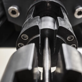 Auf dem Bild ist ein Metallstab in einer Prüfmaschine eingespannt, die für mechanische Untersuchungen von Werkstoffen eingesetzt wird. Die Aufnahme zeigt die Spannvorrichtungen der Maschine aus nächster Nähe und hebt die präzise technische Konstruktion hervor. Der Fokus liegt auf dem Prüfbereich, während die übrigen Maschinenteile im Hintergrund unscharf erscheinen.