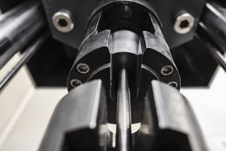 Auf dem Bild ist ein Metallstab in einer Prüfmaschine eingespannt, die für mechanische Untersuchungen von Werkstoffen eingesetzt wird. Die Aufnahme zeigt die Spannvorrichtungen der Maschine aus nächster Nähe und hebt die präzise technische Konstruktion hervor. Der Fokus liegt auf dem Prüfbereich, während die übrigen Maschinenteile im Hintergrund unscharf erscheinen.
