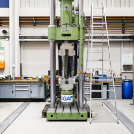Auf dem Bild ist eine große Prüfmaschine in einer Industriehalle zu sehen. Die Anlage steht frei im Raum und wird von einer danebenstehenden Leiter sowie Werkbänken und technischen Einrichtungen umgeben. Die Maschine dient der Materialprüfung und ragt mit ihrer hohen Bauweise deutlich über die umliegende Ausstattung hinaus.