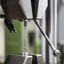 Auf dem Bild ist eine Materialprüfmaschine zu sehen, in der eine schmale Metallprobe zwischen zwei Spannbacken eingespannt ist. Die Probe befindet sich im Prüfbereich der Anlage und wird für eine mechanische Untersuchung belastet. Die Aufnahme konzentriert sich auf die präzise Prüfapparatur, während der Hintergrund unscharf dargestellt ist.