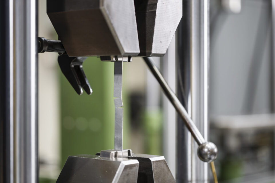 Auf dem Bild ist eine Materialprüfmaschine zu sehen, in der eine schmale Metallprobe zwischen zwei Spannbacken eingespannt ist. Die Probe befindet sich im Prüfbereich der Anlage und wird für eine mechanische Untersuchung belastet. Die Aufnahme konzentriert sich auf die präzise Prüfapparatur, während der Hintergrund unscharf dargestellt ist.