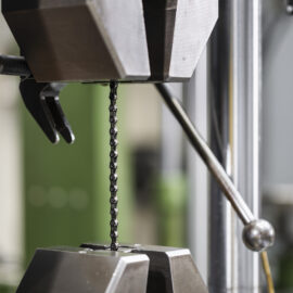 Auf dem Bild ist eine feine Metallkette in einer Materialprüfmaschine eingespannt. Die Kette wird zwischen zwei Spannbacken gehalten und für eine mechanische Belastungs- oder Zugprüfung untersucht. Der Fokus liegt auf dem Prüfbereich, während die übrigen Teile der Anlage im Hintergrund unscharf dargestellt sind.