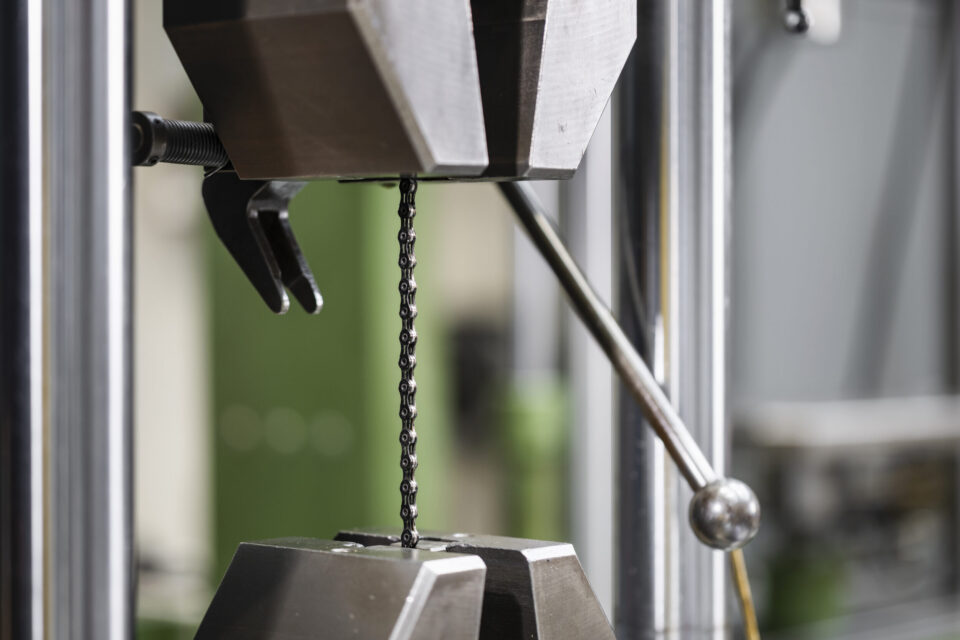 Auf dem Bild ist eine feine Metallkette in einer Materialprüfmaschine eingespannt. Die Kette wird zwischen zwei Spannbacken gehalten und für eine mechanische Belastungs- oder Zugprüfung untersucht. Der Fokus liegt auf dem Prüfbereich, während die übrigen Teile der Anlage im Hintergrund unscharf dargestellt sind.