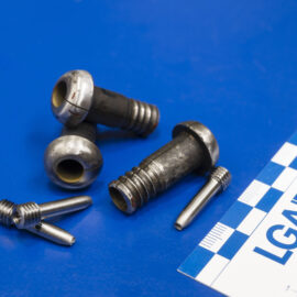 Auf dem Bild sind mehrere metallische Bauteile und Prüfproben auf einer blauen Unterlage ausgelegt. Die unterschiedlich geformten Komponenten werden im Rahmen der Materialprüfung dokumentiert und untersucht. Neben den Proben liegt ein Maßstab, der die Größenverhältnisse der Bauteile verdeutlicht.