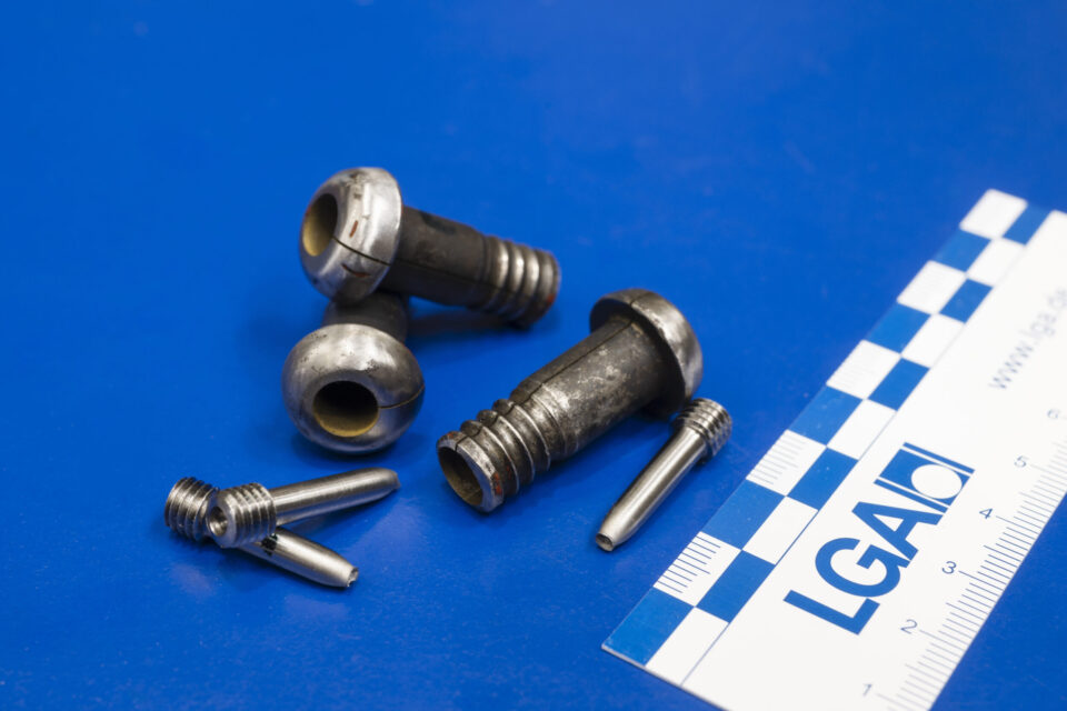 Auf dem Bild sind mehrere metallische Bauteile und Prüfproben auf einer blauen Unterlage ausgelegt. Die unterschiedlich geformten Komponenten werden im Rahmen der Materialprüfung dokumentiert und untersucht. Neben den Proben liegt ein Maßstab, der die Größenverhältnisse der Bauteile verdeutlicht.