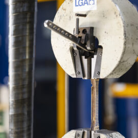 Auf dem Bild ist eine Materialprüfmaschine zu sehen, in der eine schmale Holzprobe zwischen zwei Spannvorrichtungen eingespannt ist. Die Probe wird für eine Zugprüfung belastet, um ihre mechanischen Eigenschaften und ihr Verhalten unter Beanspruchung zu untersuchen. Die Aufnahme zeigt den Prüfbereich der Maschine in Nahaufnahme und hebt die präzise Prüftechnik hervor.