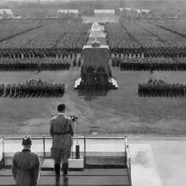 Historische Schwarz-Weiß-Aufnahme einer nationalsozialistischen Massenveranstaltung auf dem Zeppelinfeld in Nürnberg mit Adolf Hitler am Rednerpult und in Formation aufgestellten Menschen.
