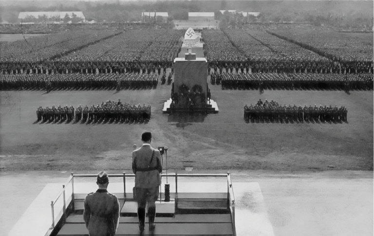 Historische Schwarz-Weiß-Aufnahme einer nationalsozialistischen Massenveranstaltung auf dem Zeppelinfeld in Nürnberg mit Adolf Hitler am Rednerpult und in Formation aufgestellten Menschen.