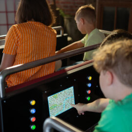 > Kinder sitzen in den Fahrzeugen der Fahrattraktion „Scotland Yard Ride“ und bedienen über integrierte Touchscreens eine digitale Karte, die während der Fahrt zur Orientierung und Interaktion dient.