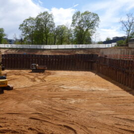 > Baugrube eines Großbauprojekts mit Spundwandverbau, Bagger und Muldenkipper während der Erdarbeiten. Die Aufnahme zeigt den vorbereiteten Baugrubenbereich für die Gründung eines neuen Gebäudes.