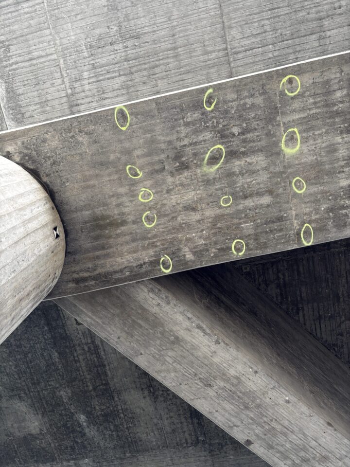Auf dem Bild ist die Unterseite eines Betonbauwerks mit einer Reihe markierter Prüfpunkte zu sehen. Die farbigen Kreismarkierungen kennzeichnen Bereiche, die im Rahmen einer Bauwerksprüfung genauer untersucht oder dokumentiert werden. Im Hintergrund sind massive Betonstützen und Tragelemente der Konstruktion erkennbar.