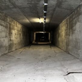 Auf dem Bild ist ein langer unterirdischer Betongang mit massiven Wänden, Decke und Boden zu sehen. Einzelne Deckenleuchten beleuchten den geradlinigen Tunnel, der in der Ferne im Dunkel verschwindet. Die Aufnahme vermittelt den Eindruck eines technischen Bauwerks oder Versorgungstunnels aus Beton.