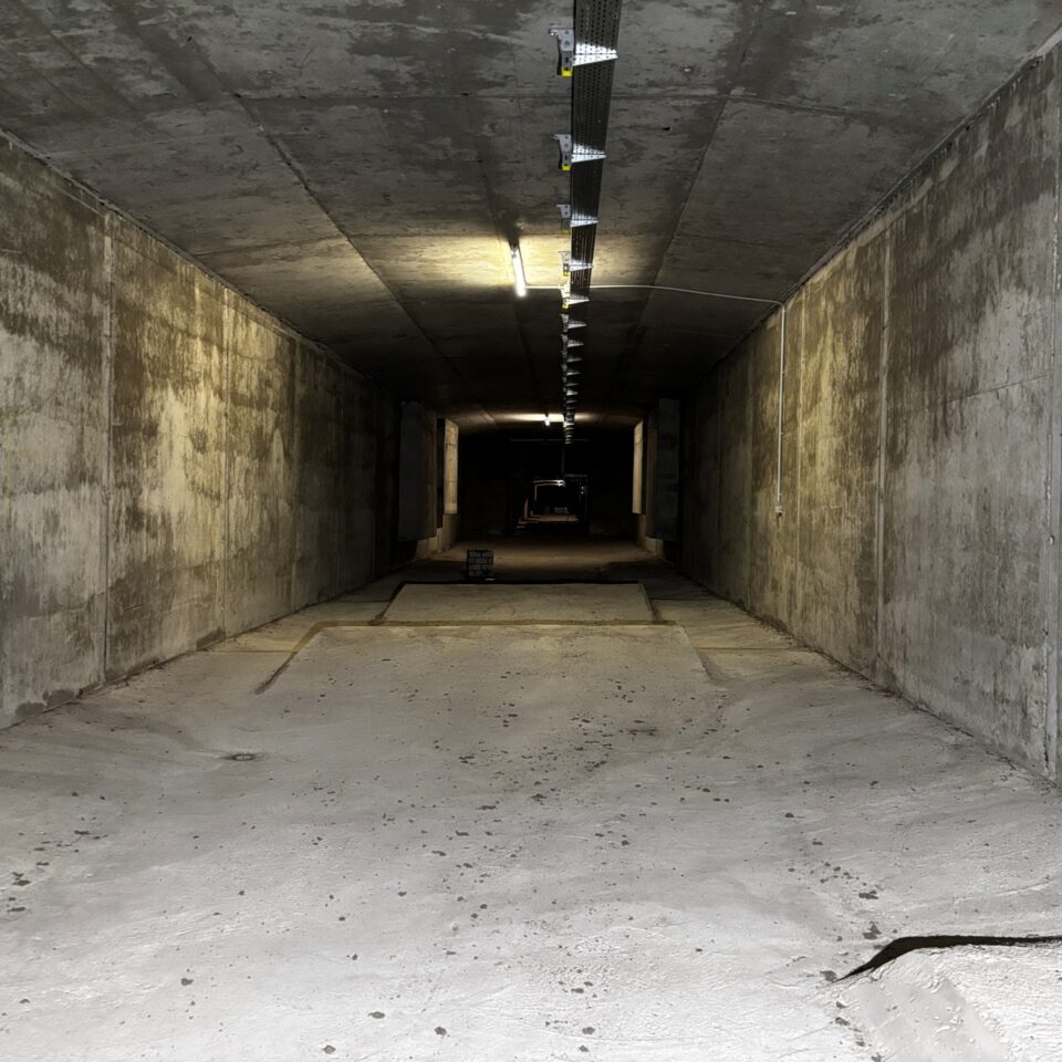 Auf dem Bild ist ein langer unterirdischer Betongang mit massiven Wänden, Decke und Boden zu sehen. Einzelne Deckenleuchten beleuchten den geradlinigen Tunnel, der in der Ferne im Dunkel verschwindet. Die Aufnahme vermittelt den Eindruck eines technischen Bauwerks oder Versorgungstunnels aus Beton.