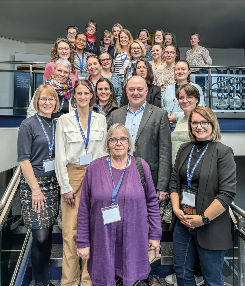 Auf dem Bild ist eine große Gruppe von Personen auf einer Treppe in einem Gebäude zu sehen, die gemeinsam für ein Gruppenfoto posieren. Die Teilnehmenden tragen Namensschilder und blicken lächelnd in die Kamera. Das helle Treppenhaus mit Geländern und offenen Ebenen bildet den Hintergrund der Aufnahme.