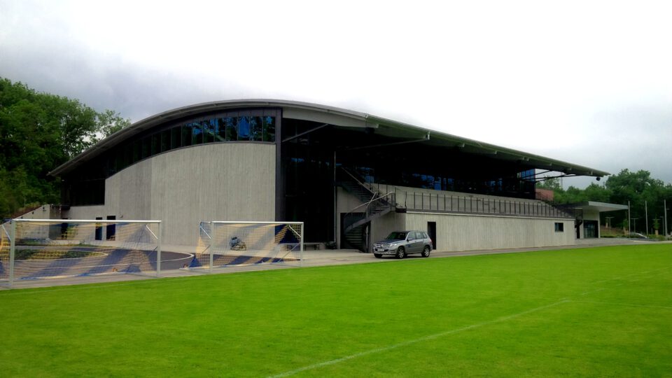 Moderne Sportanlage mit geschwungenem Dach und großen Glasfenstern, davor ein gepflegter Rasen mit zwei Fußballtoren und ein geparktes silbernes Auto nahe dem Eingang.