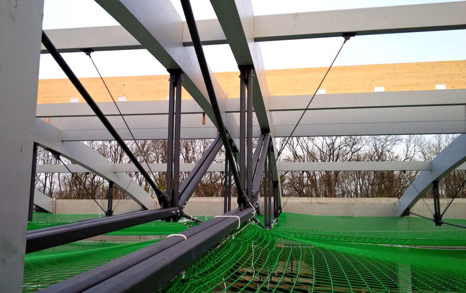 Metallkonstruktion mit Trägern und Sicherungsseilen, darunter ein grünes Sicherheitsnetz; im Hintergrund kahle Bäume und klarer Himmel.