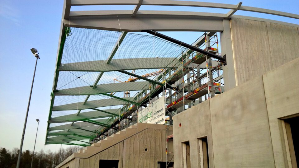 Baustelle mit teilweise errichteter Metallkonstruktion, Dach mit Netzsystem, umgeben von Gerüsten.