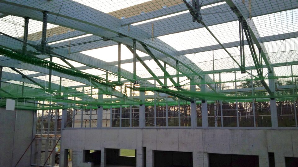 Innenansicht eines Rohbaus mit Betonwänden, großen Fenstern und einem grünen Sicherheitsnetz unter dem teilweise offenen Dach, getragen von Metallträgern.