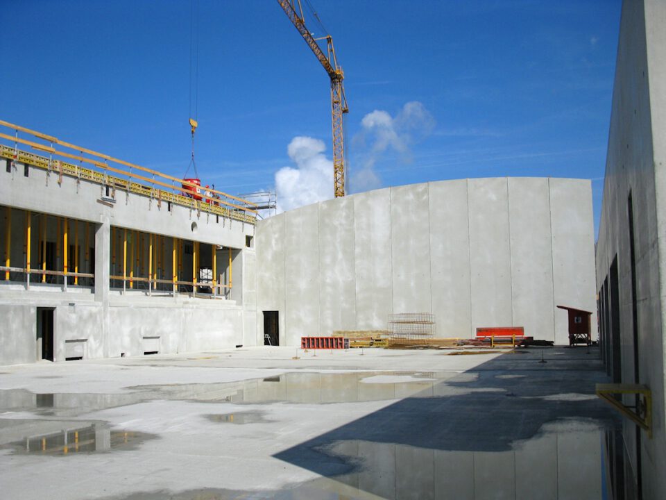 Baustelle mit großen Betonwänden und gelben Geländern, im Hintergrund ein Kran; auf dem betonierten Boden befinden sich Wasserpfützen und Baumaterialien.