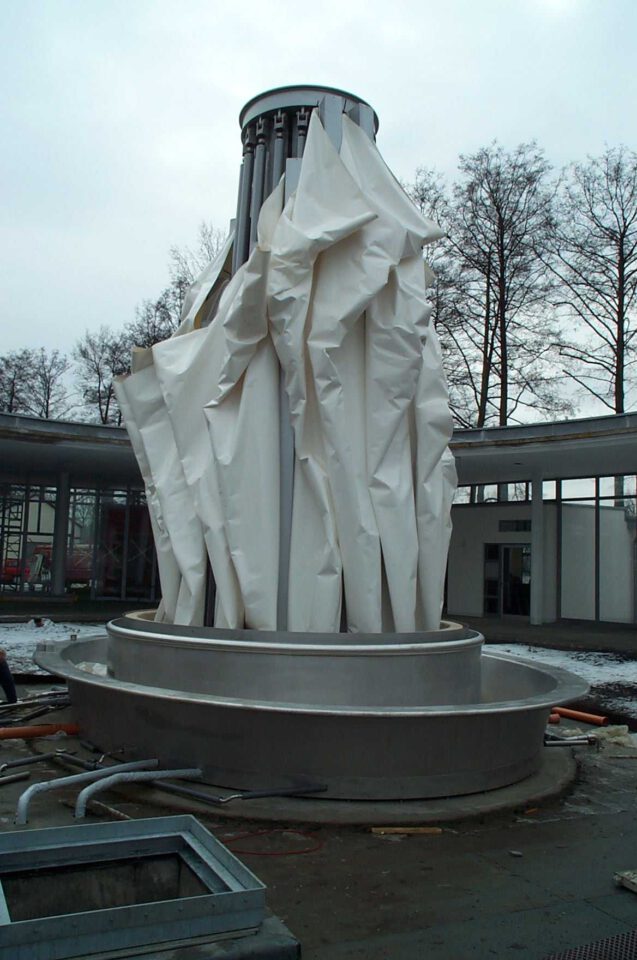 Große zylindrische Struktur mit metallischem Sockel und oberem Teil aus zerknittertem weißem Material, im Freien zwischen Bäumen und Gebäuden; am Boden liegen Rohre und Baumaterialien.