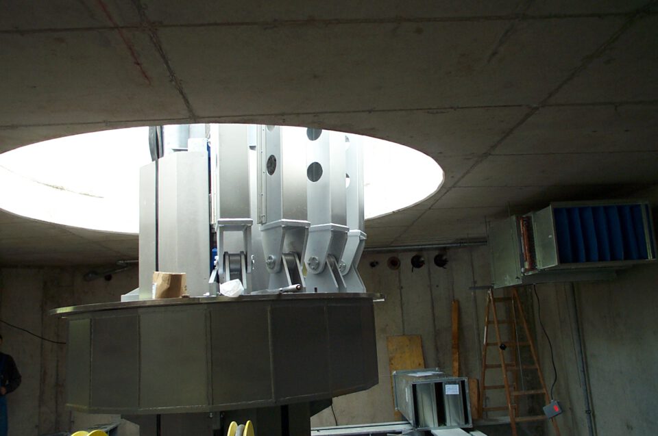 Industrieller Innenraum mit Betonwänden und kreisförmiger Öffnung in der Decke; im Zentrum eine große metallische Anlage, rechts eine Holzleiter und weiteres technisches Gerät.