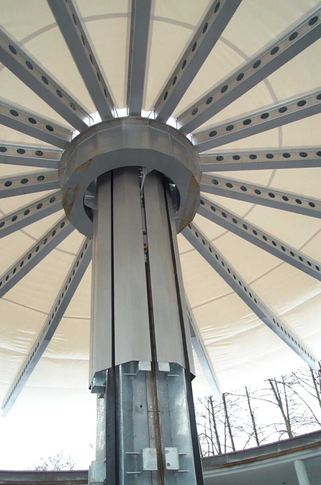 Innenansicht einer großen, runden Überdachung mit zentraler Metallsäule und radial verlaufenden Trägern; darüber ist ein helles Stoffdach gespannt.