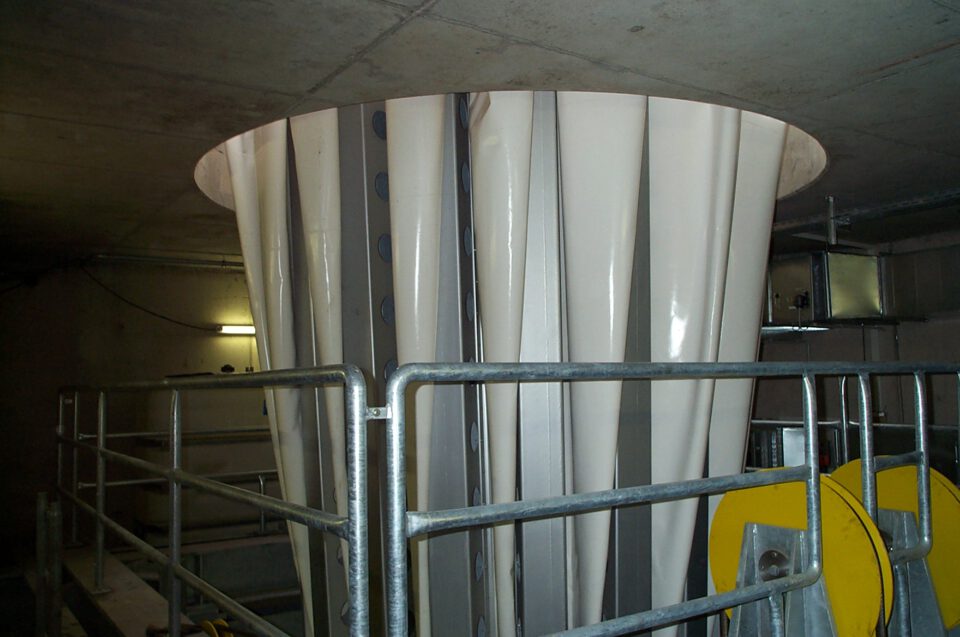 Industrielle Anlage mit großem, weißem Kegelbau aus Stoff und Metallstreben, umgeben von Geländern; rechts gelbe mechanische Bauteile, umgeben von Betonwänden und -decke.