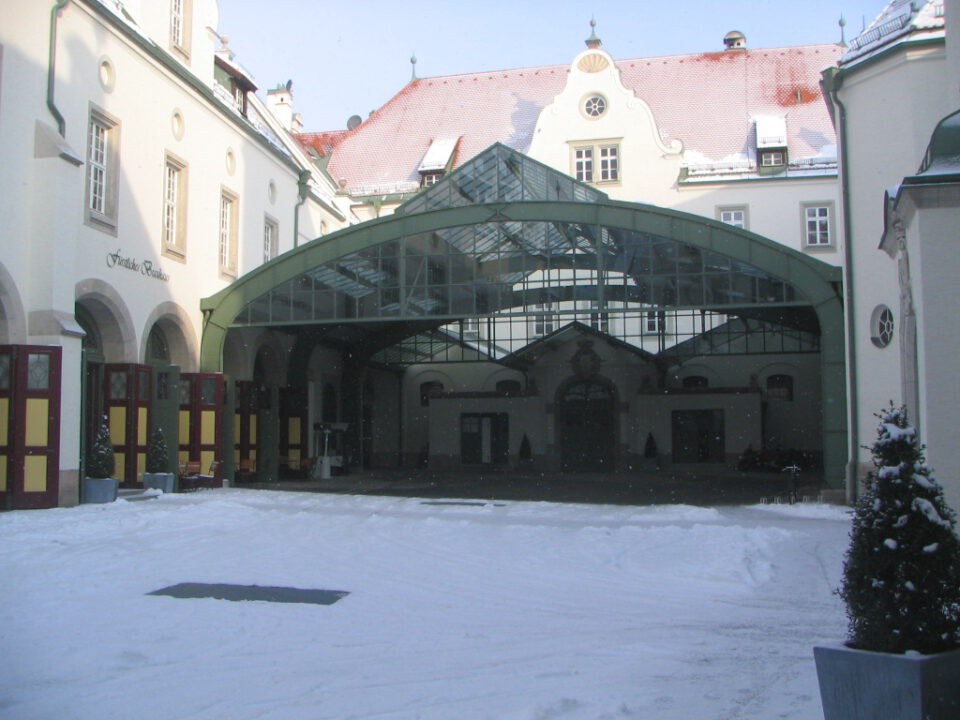 Innenhof eines Gebäudekomplexes mit europäischer Architektur, verschneitem Boden und Dächern, sowie einer großen Glasüberdachung über einem Teil des Hofes, was auf Winterzeit hinweist.