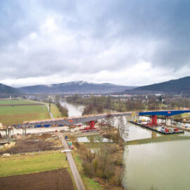Baustelle an einem Fluss mit einer im Bau befindlichen Brücke; sichtbar sind Kräne, Baumaschinen und Materialien, umgeben von Feldern, Bäumen und Hügeln im Hintergrund.