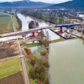 Brückenbaustelle über einem Fluss in ländlicher Umgebung, mit sichtbaren Baumaschinen und Materialien auf beiden Uferseiten; umgeben von Feldern, Straßen, Hügeln und einer Ortschaft im Hintergrund, aufgenommen aus erhöhter Perspektive.