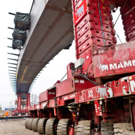 Großes, rotes Schwerlasttransportfahrzeug mit zahlreichen Rädern, das ein großes Brückensegment befördert.