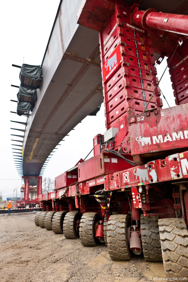 Großes, rotes Schwerlasttransportfahrzeug mit zahlreichen Rädern, das ein großes Brückensegment befördert.
