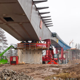 Brückenbaustelle mit massiven Betonpfeilern, mehreren Baumaschinen wie einem grünen und einem rotem Kran; das umliegende Gelände ist schlammig mit Erdhaufen und Gestein.