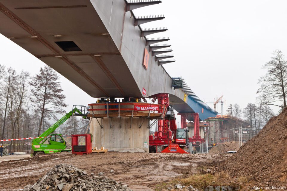 Brückenbaustelle mit massiven Betonpfeilern, mehreren Baumaschinen wie einem grünen und einem rotem Kran; das umliegende Gelände ist schlammig mit Erdhaufen und Gestein.