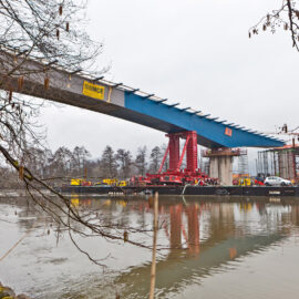 Brückenbaustelle über einem Gewässer mit teilweise montierten blauen und grauen Brückensegmenten; Kräne, Baumaschinen und Arbeiter sind sichtbar, eingerahmt von Bäumen und Ästen in natürlicher Umgebung.